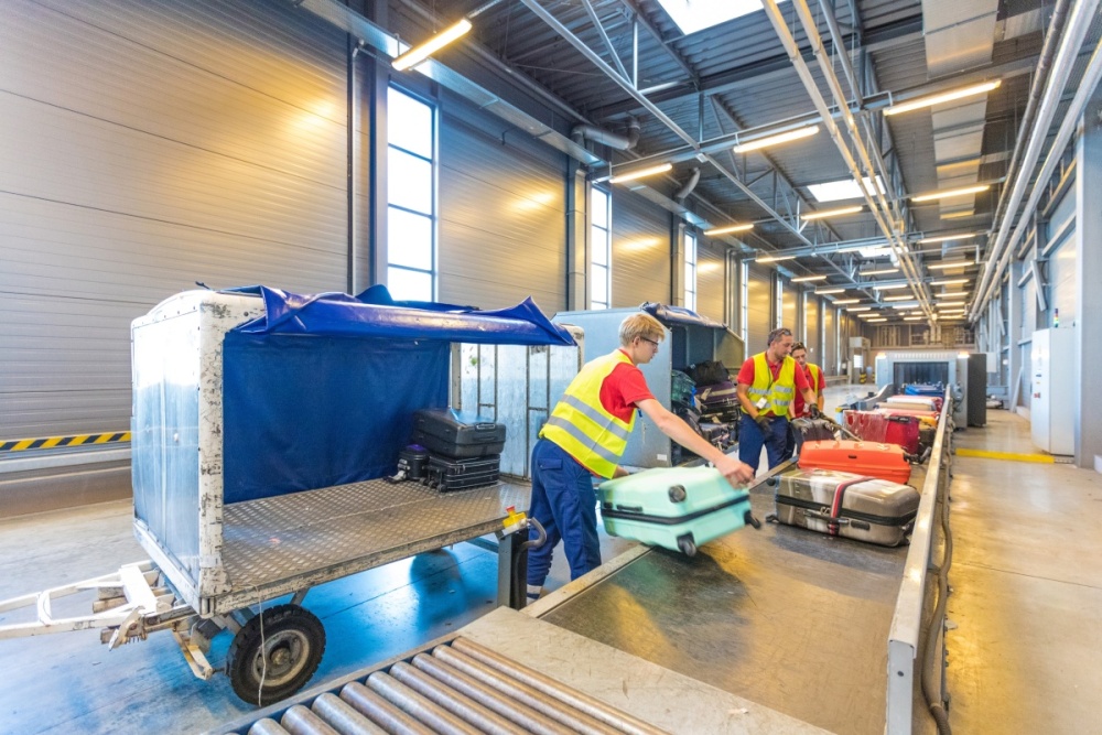 Airport employees move baggage from a conveyor belt to a baggage cart.