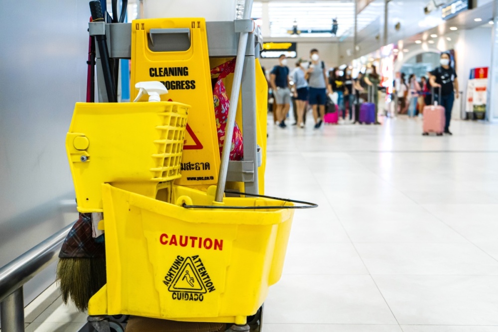 A janitorial cart with bucket and mop sit in the aisle of a busy airport.