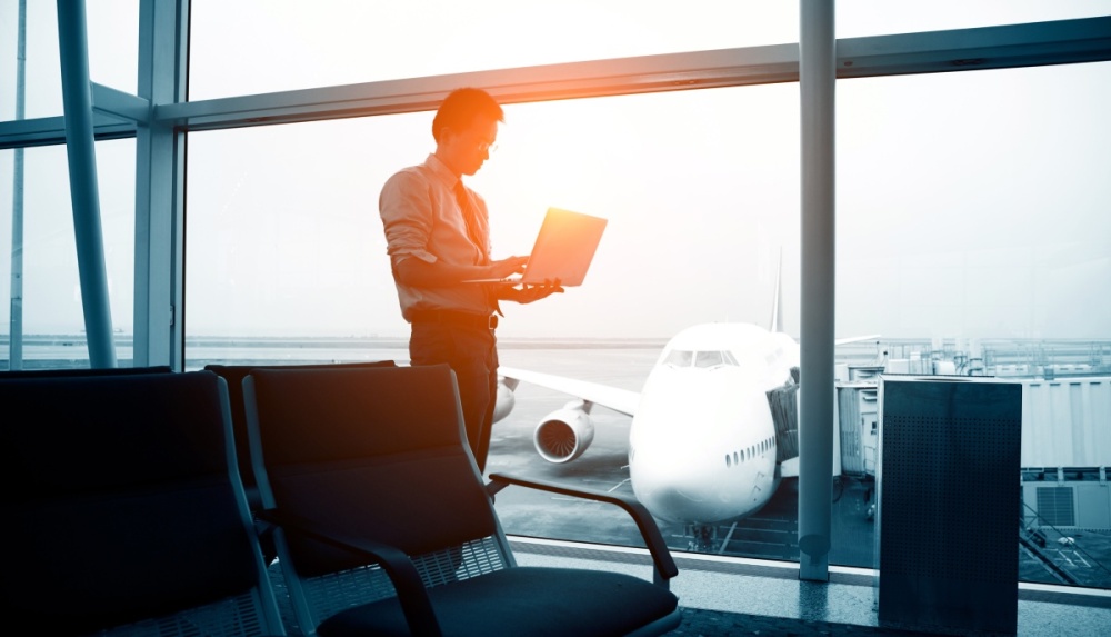 A man with a laptop is standing in the window of an airport terminal. Outside the window, a plane sits at the gate.