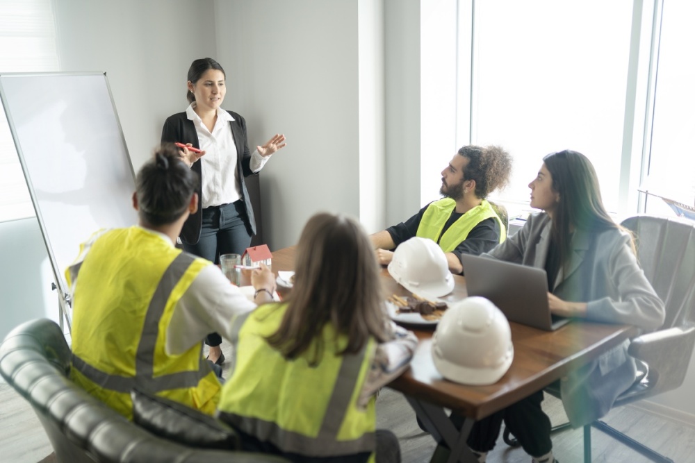 A woman at a whiteboard is presenting to four young professionals who are sitting at a table.